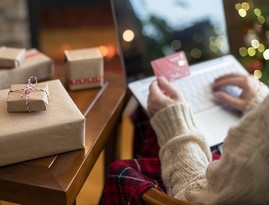 Christmas. Woman in sweater holding credit card using laptop for making order sitting at table with packaging gift near fireplace and christmas tree. Online shopping concept